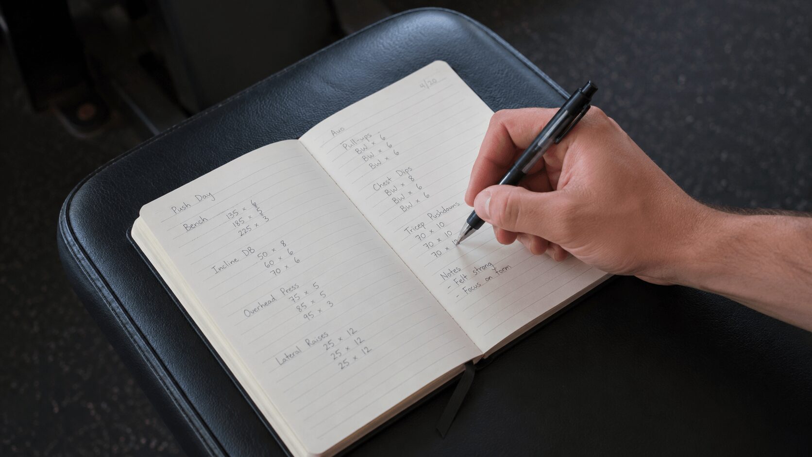ジムでトレーニングノートに記録をつけている男性の手元。ペンで数字を書いている