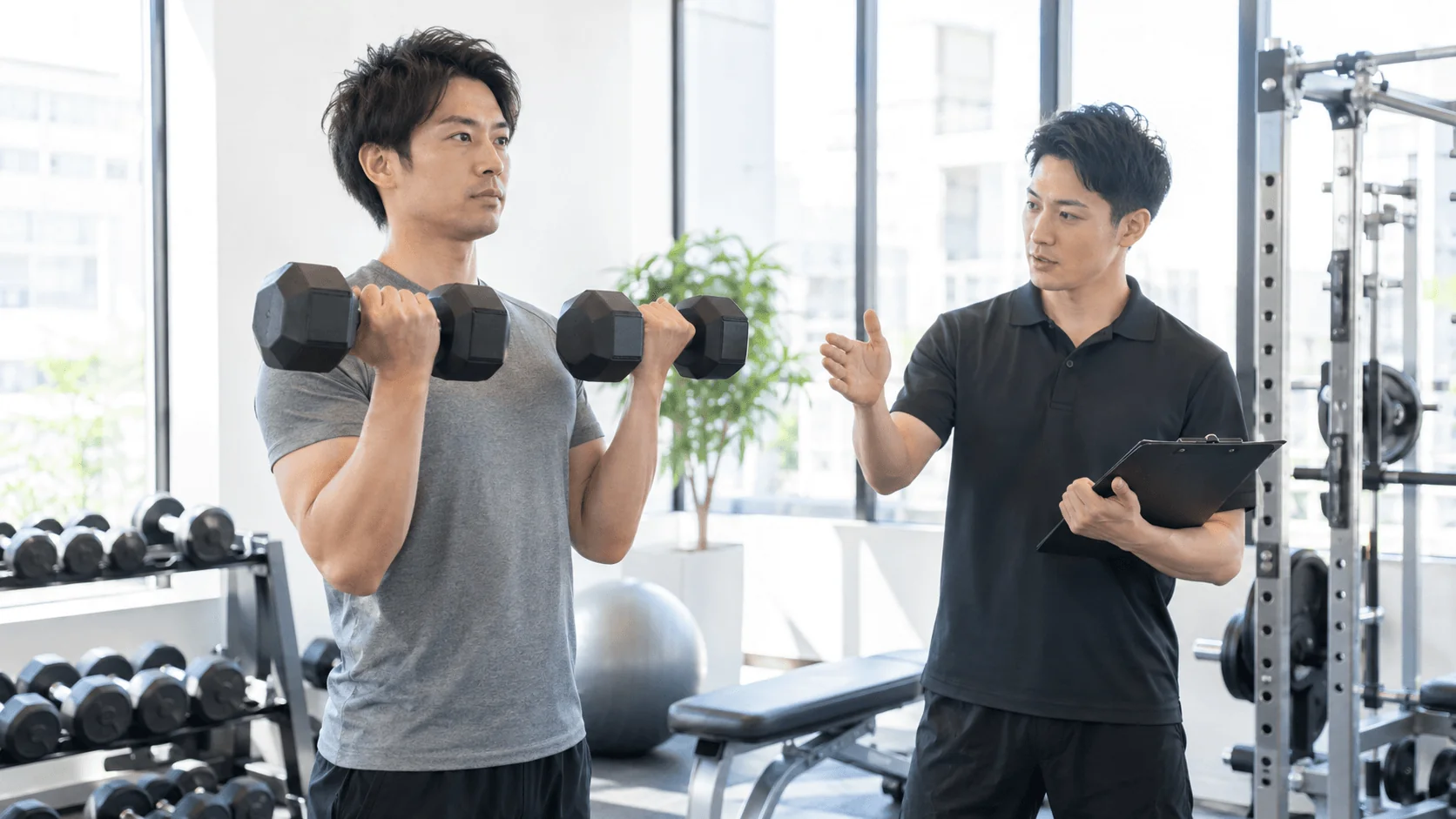トレーナーがクリップボードを見ながら男性の筋トレを管理している様子